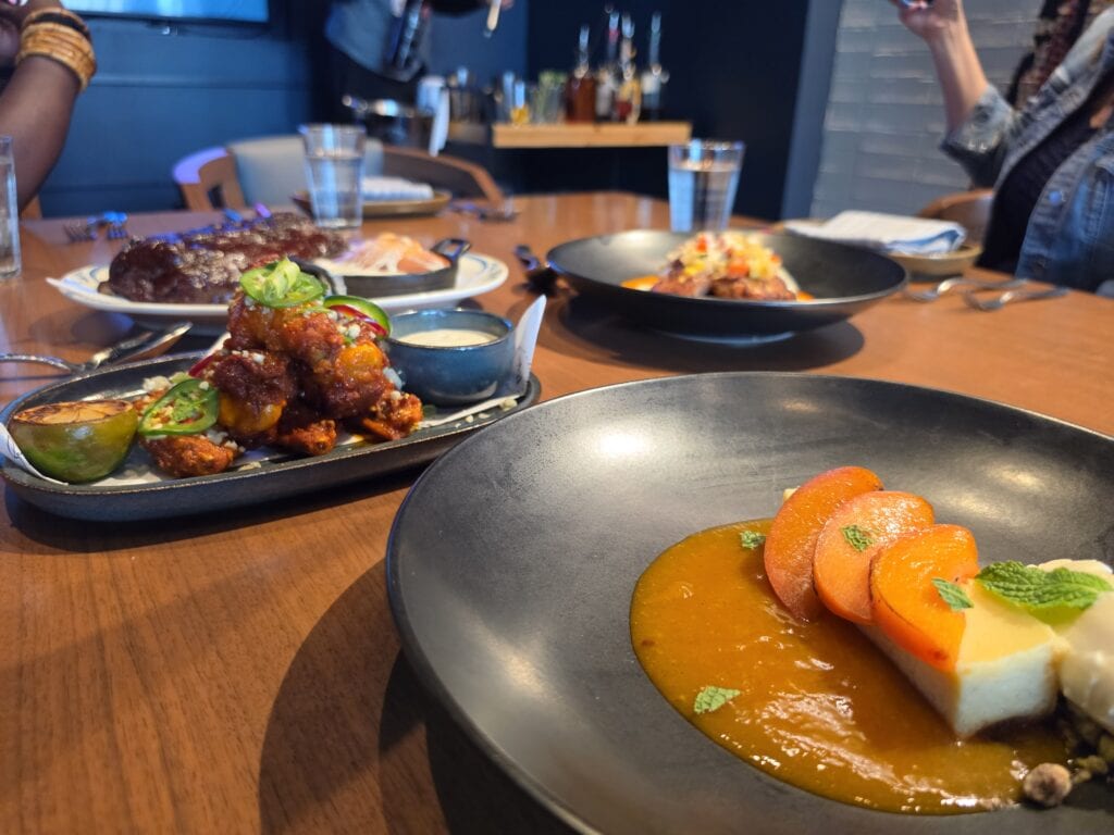 A table at Ramsays Kitchen inside Caesars Virginia in Danville VA is covered with several plated dishes including glazed fried chicken topped with sliced jalapenos and green onions. A large black plate in the foreground holds a dessert with bright orange fruit and sauce while steak and other entrees fill the table behind it.