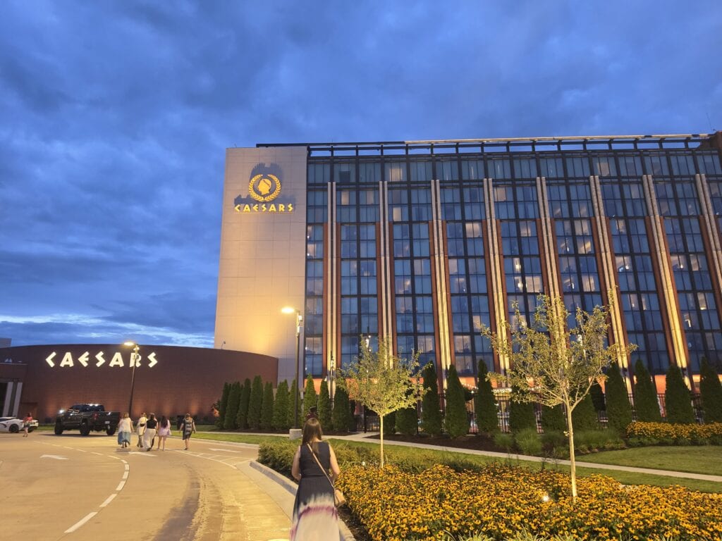 Caesars Virginia in Danville VA glows at dusk with a tall glass hotel tower and illuminated "CAESARS" signs on the building exterior. Guests walk toward the entrance past landscaped flower beds and young trees under a deep blue cloudy sky making Caesars Virginia one of the prominent things to do in Danville VA.