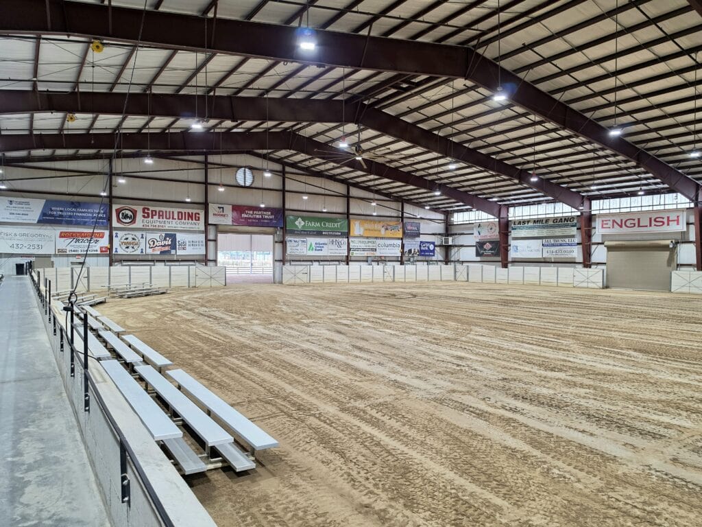 The indoor arena at the Olde Dominion Agricultural Complex in Chatham, VA has a large dirt floor surrounded by white fencing and rows of metal bleachers beneath a high exposed beam ceiling. Sponsor signs line the far wall including "Spaulding Equipment Company" and "Farm Credit" while larger signs read "Last Mile Gang" and "English". The spacious covered arena hosts agricultural and equestrian events and offers another option for things to do in Danville VA.