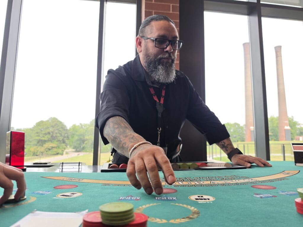 A blackjack dealer at Caesars Virginia in Danville VA reaches across a green felt table while teaching players how to play. Colorful casino chips sit near the edge of the table with large windows and brick smokestacks visible behind the dealer.