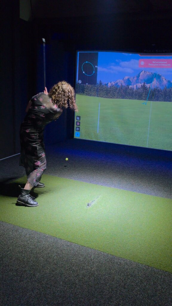 Phoenyx swings a golf club toward a ball on a green hitting mat inside the virtual golf simulator at River District Social in Danville VA. A large projection screen shows a digital golf course and the message "Ball not detected. Please place on hitting mat." as part of the interactive game.