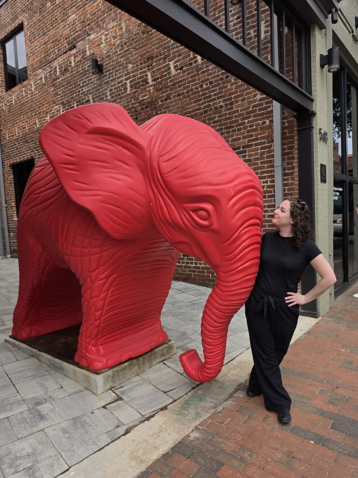 Phoenyx poses beside a massive bright red elephant sculpture during the SoSi Safari in downtown Danville VA. She stands near the elephants lowered trunk on a brick sidewalk creating a playful stop while exploring things to do in Danville VA.