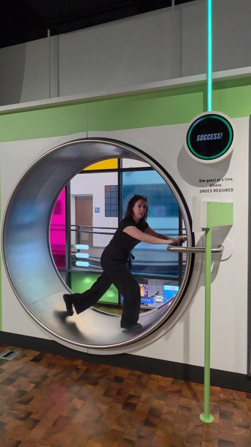 Phoenyx runs inside a giant metal wheel at the Danville Science Center as an interactive exhibit measures her movement and speed. A circular display above her reads "SUCCESS." and nearby instructions read "One guest at a time please." and "SHOES REQUIRED.".
