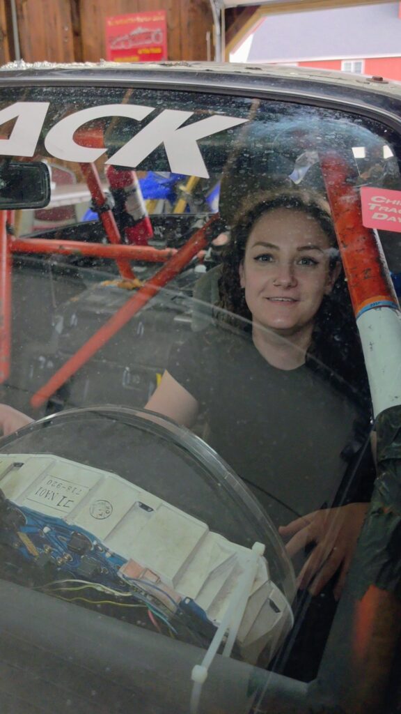 Phoenyx sits behind the wheel of a race car at Virginia International Raceway near Danville VA with a red roll cage surrounding the drivers seat. She smiles through the windshield while exploring the interior controls of the track prepared car.