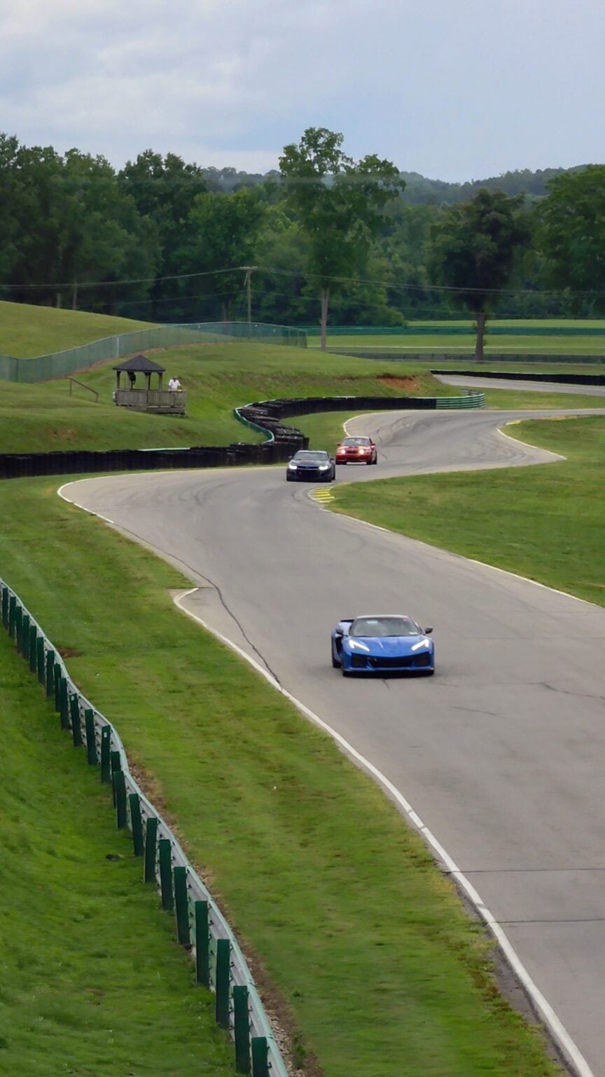 A blue sports car leads two other cars through a sweeping curve at Virginia International Raceway near Danville VA. The paved track winds through rolling green hills and trees showing the high speed racing experience available among things to do in Danville VA.