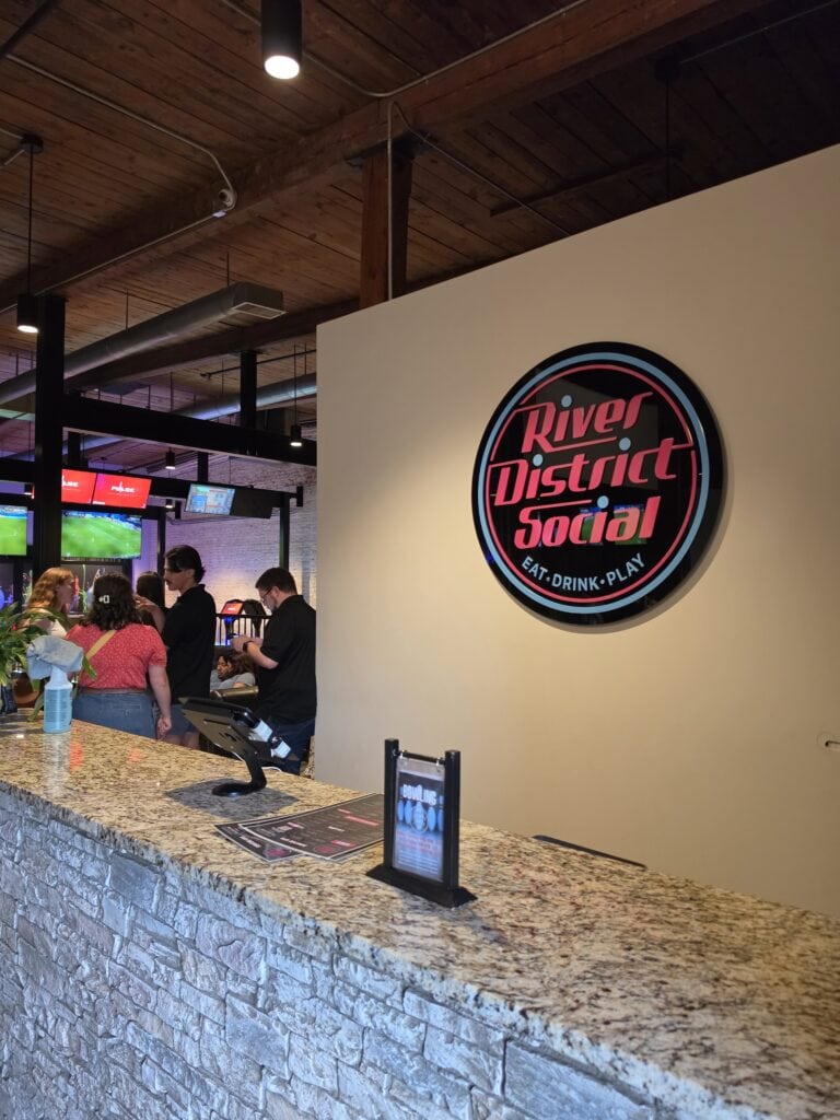 The entrance counter at River District Social in Danville VA sits beneath a large round illuminated sign reading "River District Social" and "EAT DRINK PLAY". Guests gather near arcade and sports screens in the open entertainment venue making it one of the indoor things to do in Danville VA.