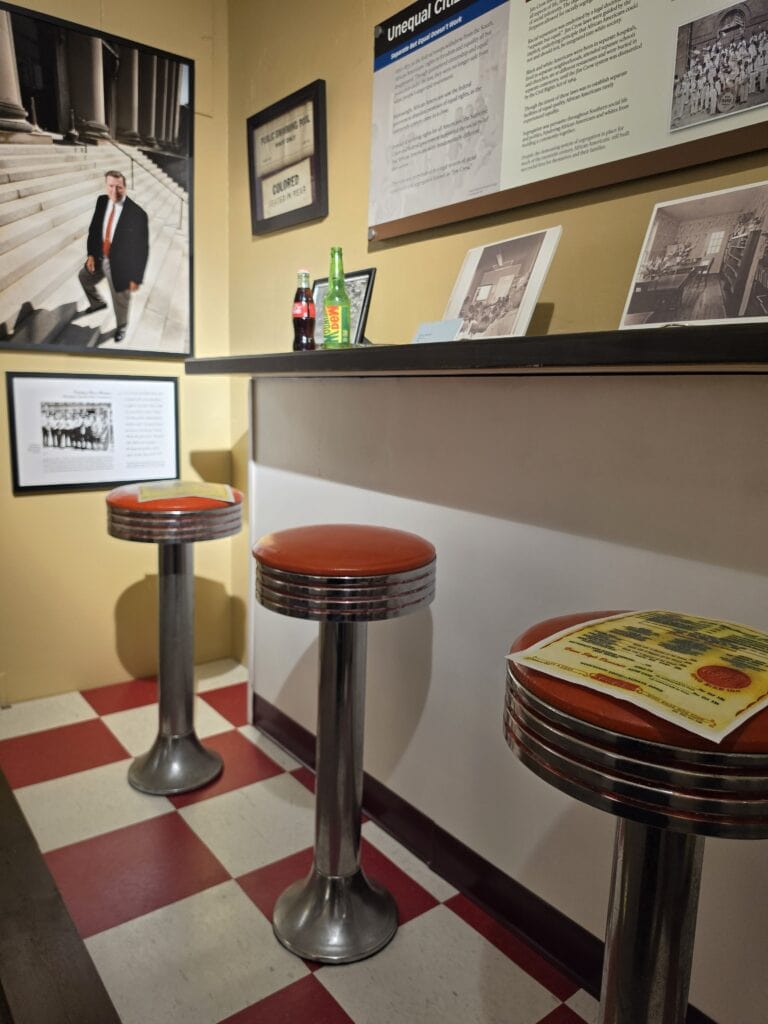 A recreated civil rights era lunch counter at the Danville Museum of Fine Arts and History features red vinyl stools chrome bases and a red and white checkered floor. Historic photographs documents and an exhibit panel titled "Unequal Cities" surround the counter to interpret the history of segregation and sit in protests in Danville.